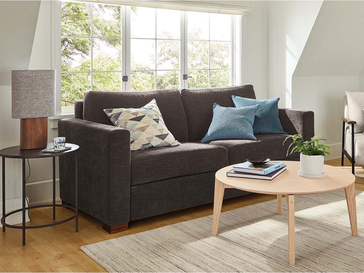 Charcoal Berin sleeper sofa with geometric and blue pillows, round wood coffee table, and windowed seating area with side table and lamp.