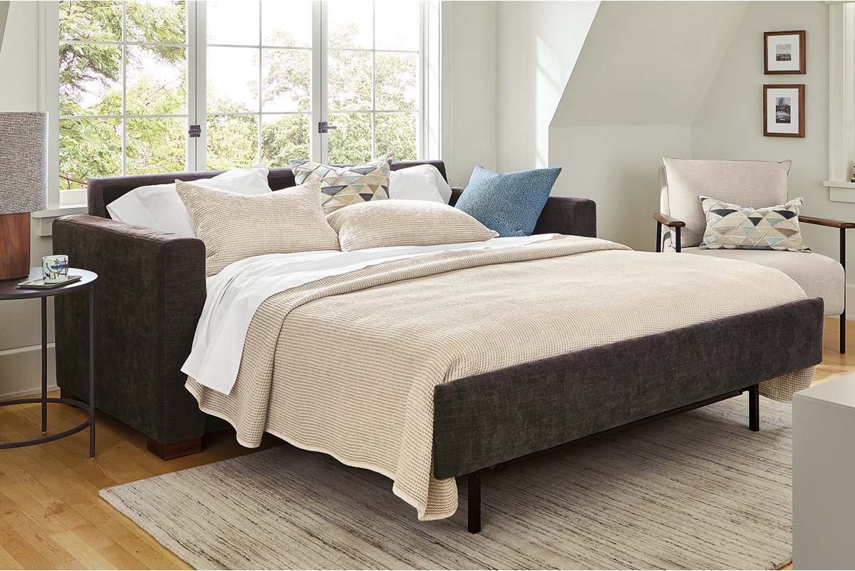 Berin sleeper sofa opened into a bed with textured bedding, layered pillows, lounge chair, side table, and framed artwork on the far wall.