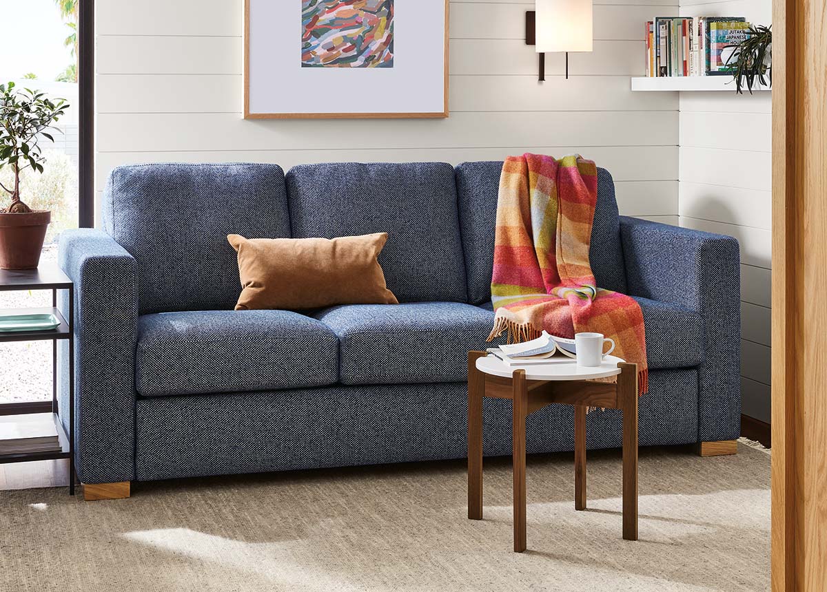 Berin sleeper sofa in textured blue fabric, layered with throw and pillow, paired with wood side table and abstract framed art.