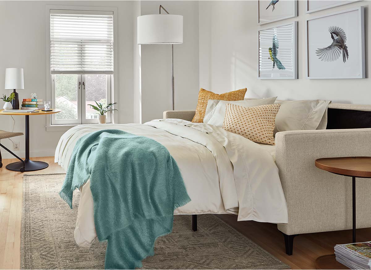 Extended Berin sleeper sofa styled with layered bedding, teal blanket, patterned pillows, side tables, and framed bird prints.