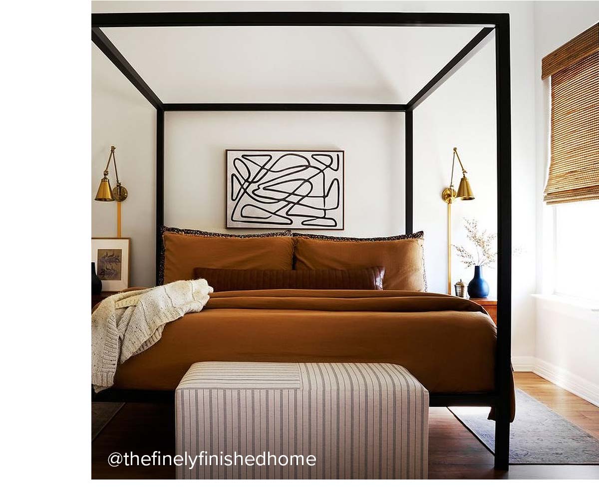 Black canopy bed with rust bedding, striped bench, and abstract art over headboard.