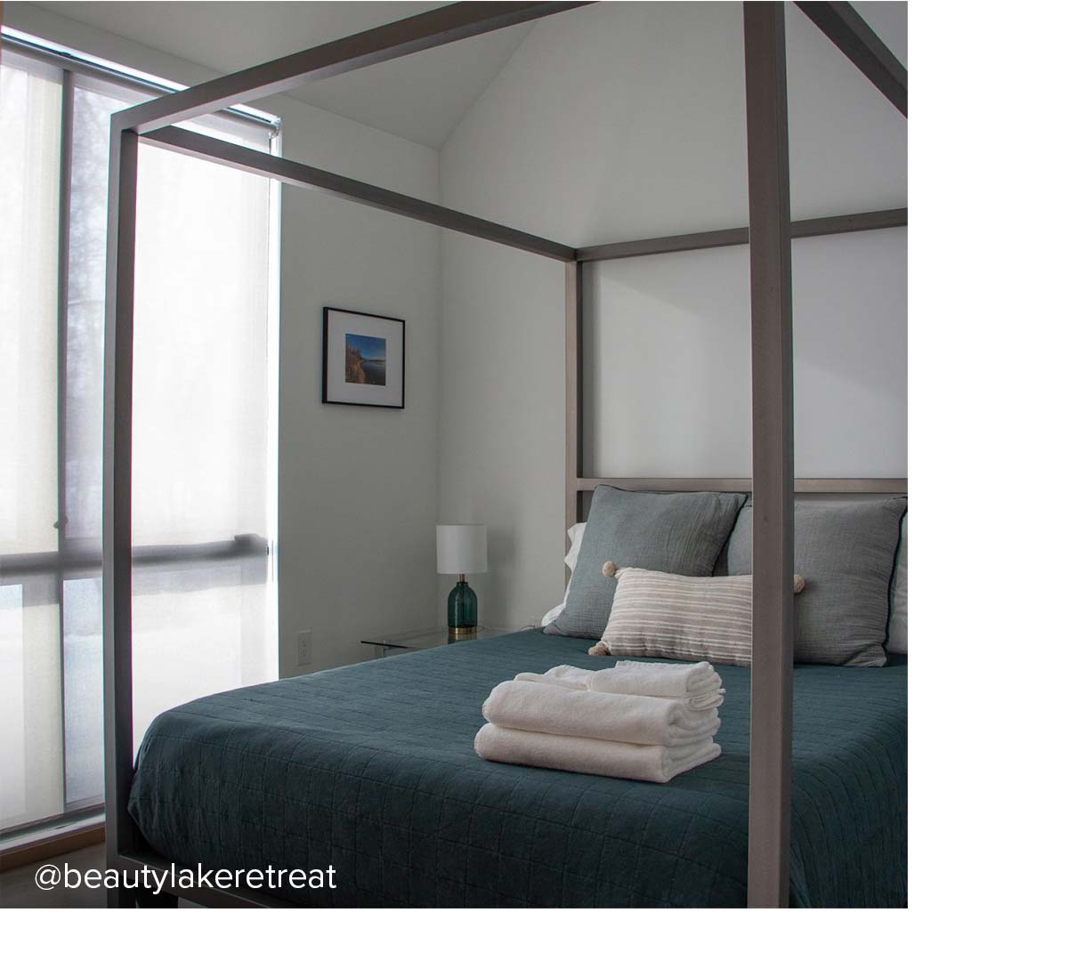 Canopy bed with grey posts, dark bedding, and large square pillows near a windowed corner.