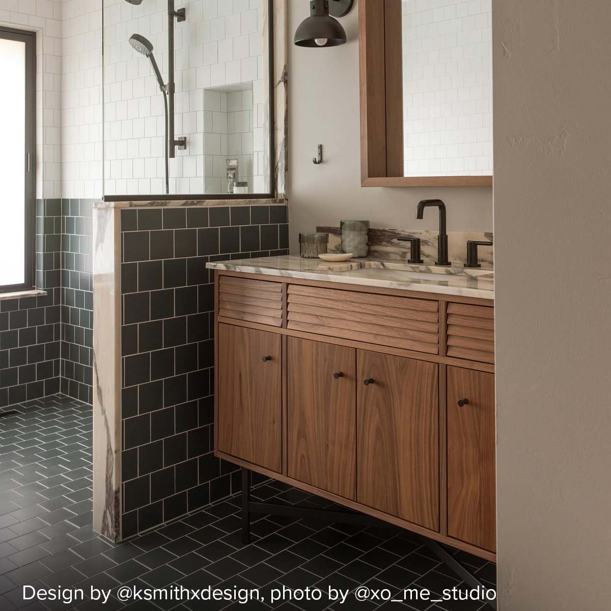 Bathroom with wood vanity, fluted drawer fronts, marble counter, and black fixtures beside tiled walk-in shower.