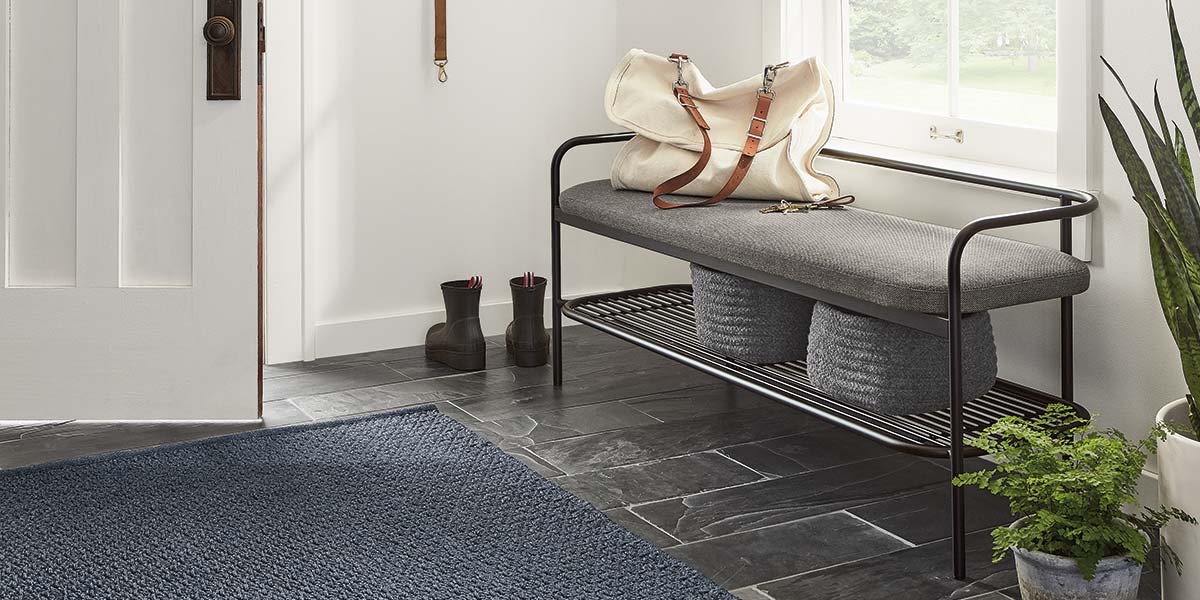 Entryway bench with a gray cushion and black metal frame, styled with a canvas bag and baskets underneath on slate flooring.