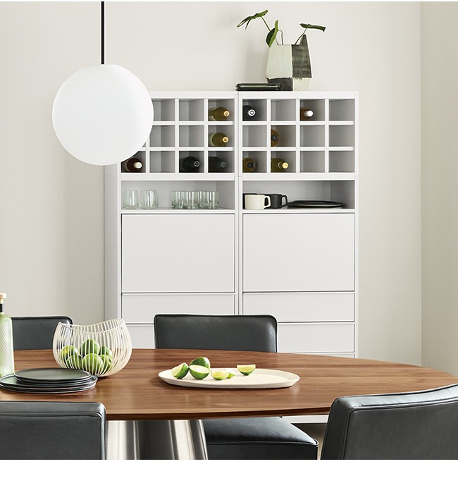 White storage system with bottle grid, drawer storage, and open shelving, placed behind round Decker table.