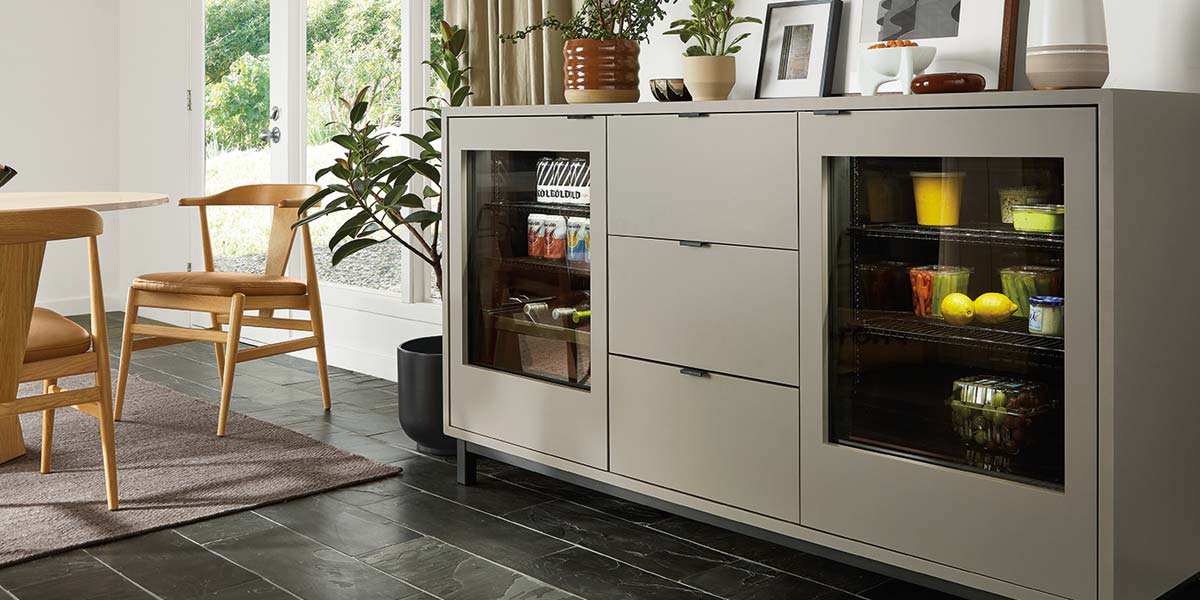 Grey storage cabinet with glass fridge doors holding drinks and produce near wood dining set.