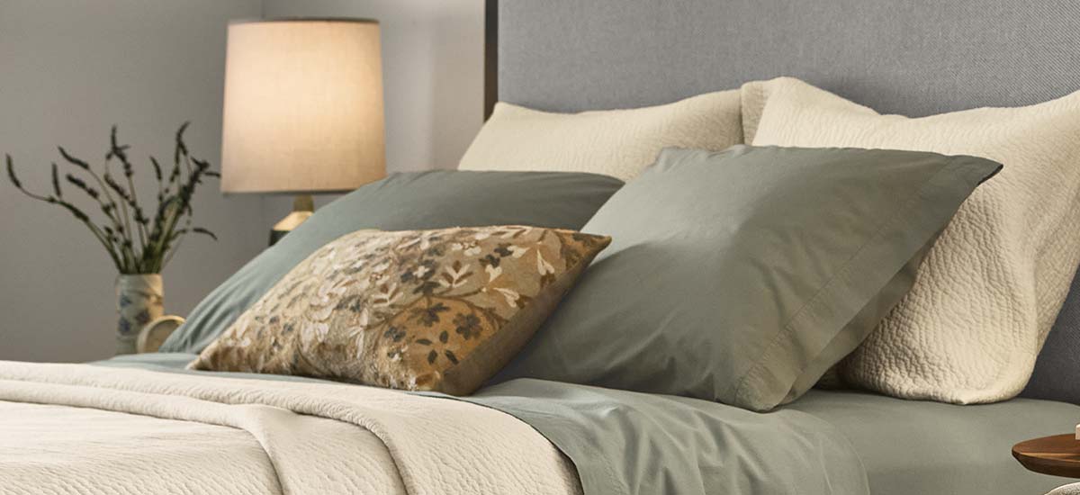 Gray upholstered headboard with layered bedding in soft green and cream tones, accented by a floral pillow.