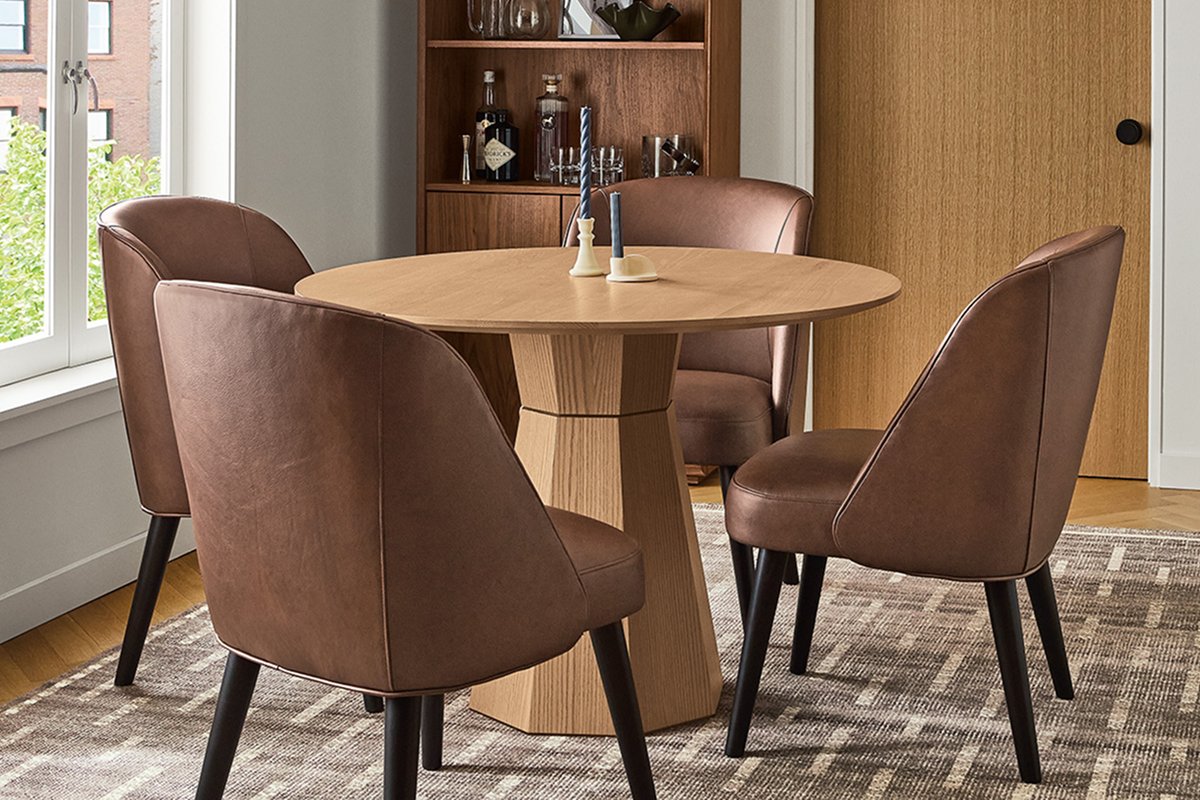 Round Glover pedestal table in light oak paired with four brown leather chairs featuring curved backs and tapered legs.