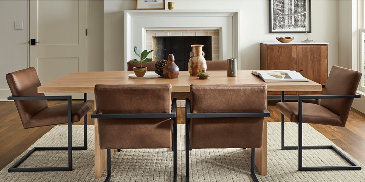 Light wood dining table with black-framed leather chairs in a neutral-toned room with fireplace and sideboard.