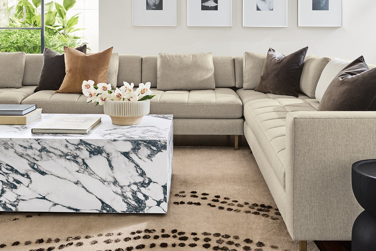 Lloyd sectional in beige fabric with marbled Leona coffee table holding books and a ribbed planter with white flowers.