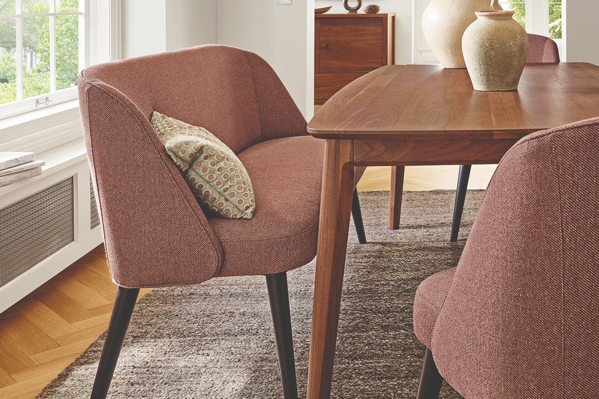 Rust-toned Cora bench with rounded back at walnut dining table on woven rug.