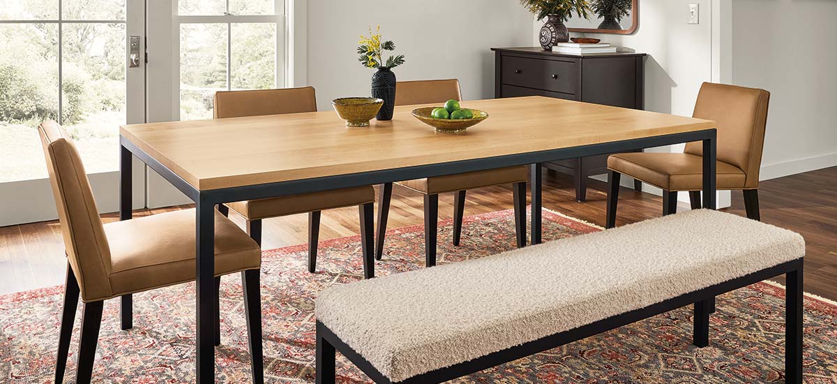 Light-top dining table with black legs paired with camel dining chairs and soft bench in bright room.