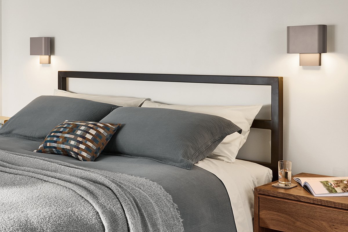 Kalli wall sconces in graphite frame, a minimalist bed with grey bedding, wood nightstand and patterned accent pillow.