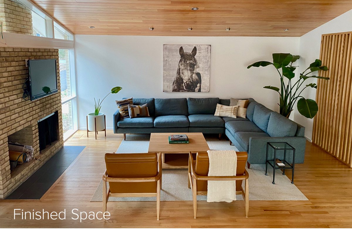 Real version of the living room with green sectional, tan lounge chairs, wood coffee table, and large plant next to artwork of horse.