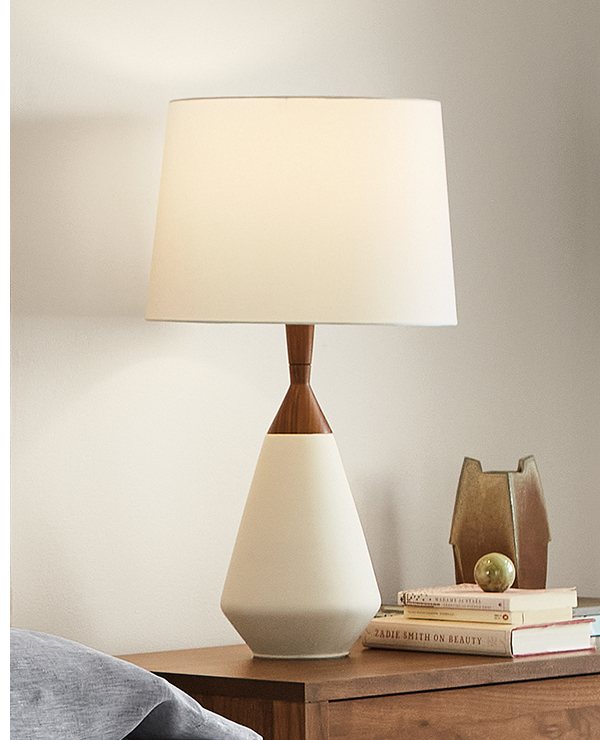 Cloak table lamp with white ceramic base, wood accent neck, and white shade on a wood nightstand.