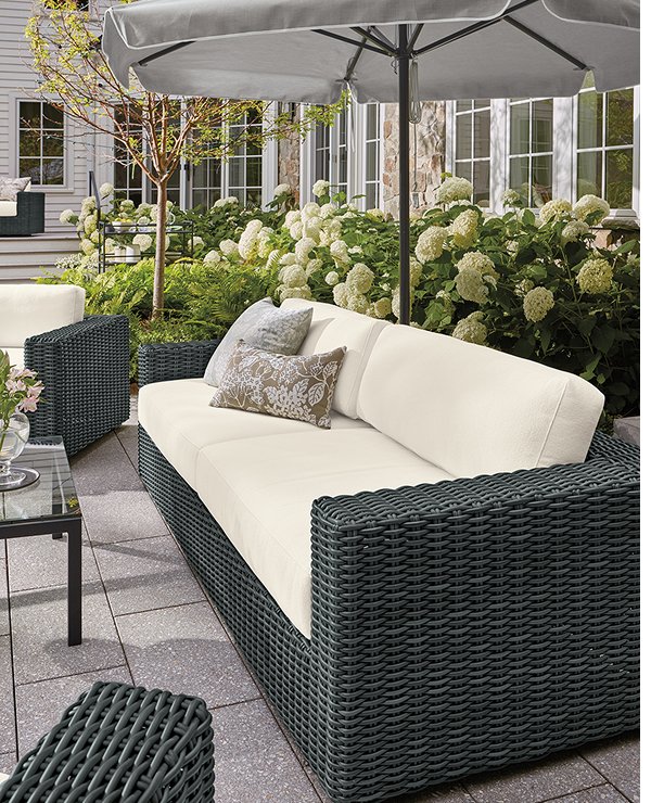 Madura outdoor sofa with dark wicker frame and white cushions, styled with patterned pillows under a patio umbrella.