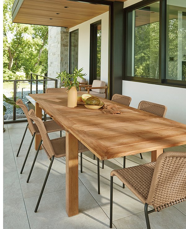 Capri outdoor dining table in teak with woven dining chairs, styled with a vase and bowls on a covered patio.