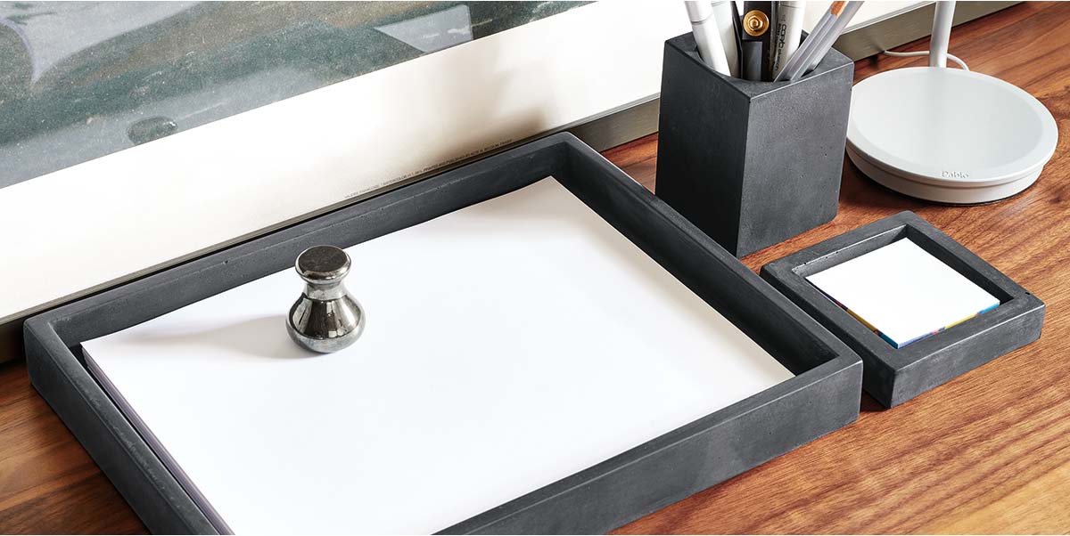 Black Saco desktop organizers with a tray, pen holder, and note holder on a wooden desk, styled with office supplies.