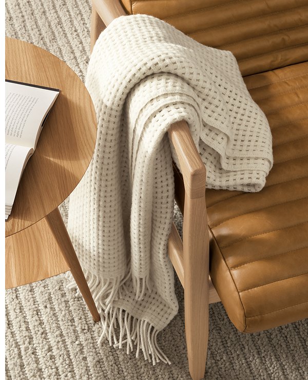 Homage throw in cream waffle knit draped over the arm of a leather Callan chair beside a table.
