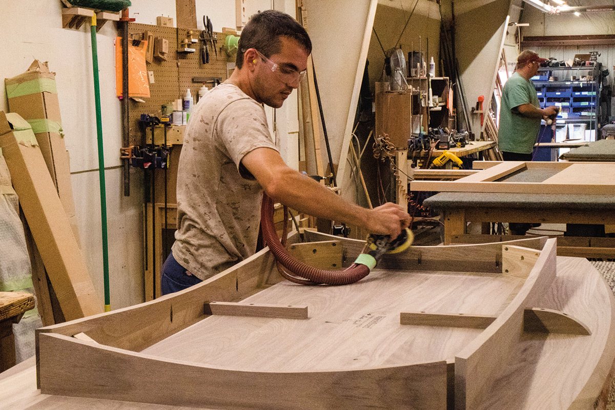 Person using a power sander on a wood table base in a workshop with tools and materials along the walls.