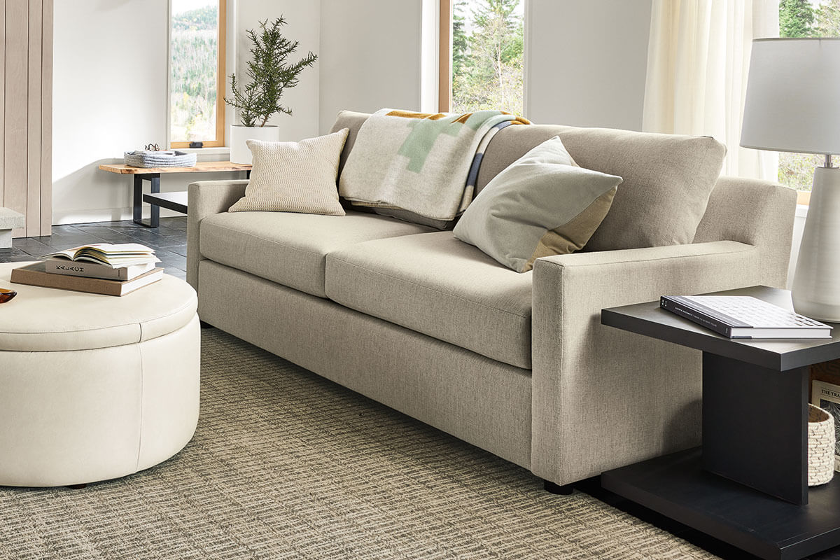 Ludlow sofa in beige fabric with plush cushions, throw blanket, and round ottoman in front.