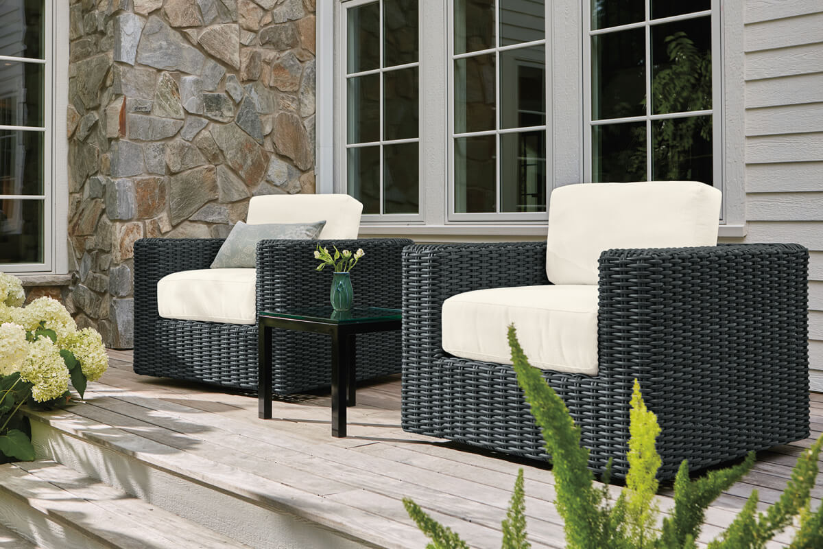 Two black wicker Madura chairs with ivory cushions sit on a wooden deck with a small glass-top side table.