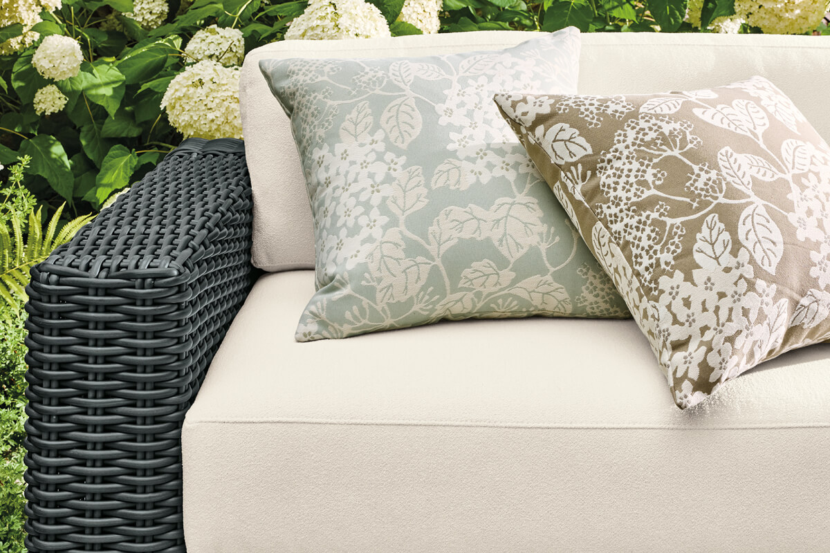 Close-up of black wicker Madura sofa arm with ivory cushions and two floral throw pillows in sage green and taupe.