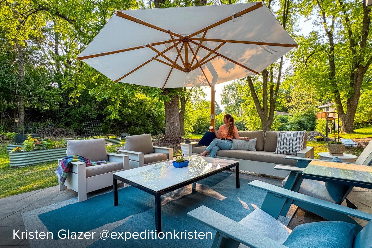 Rayo seating arranged with sofa and lounge chairs around a glass-top table under a large umbrella in a backyard patio.
