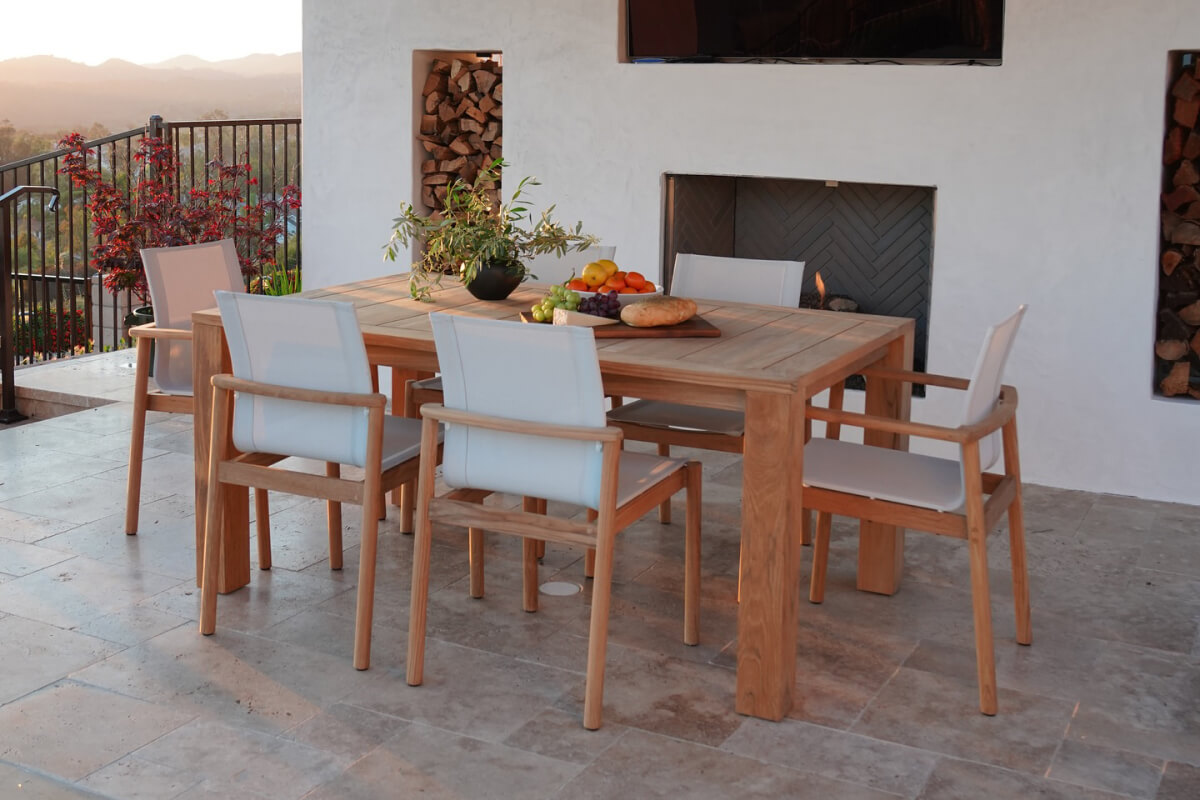 Teak Capri table with matching chairs on a patio near a fireplace, styled with fruit, bread, and a potted plant.