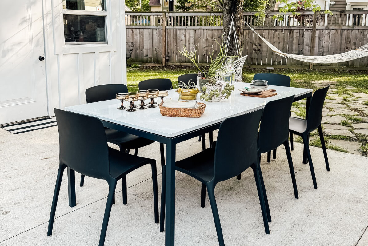 White stone top Parsons table with black chairs on a patio, styled with glassware and a centerpiece of plants and serving pieces.