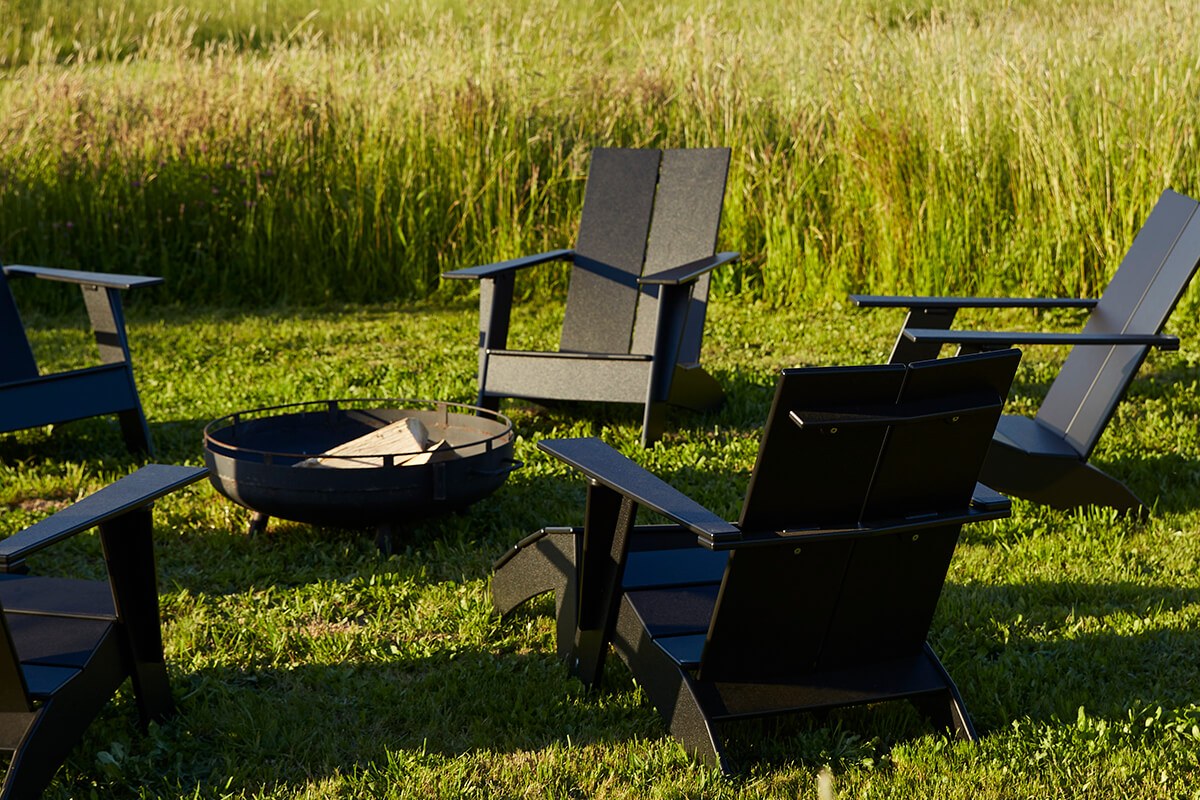 Black Emmet lounge chairs gathered around a fire pit on grass, set in an open yard with tall greenery in the background.