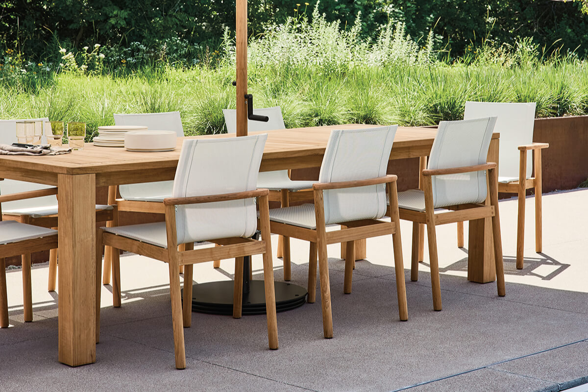Capri outdoor dining table with wood chairs and white cushions arranged on a patio beside tall grasses.