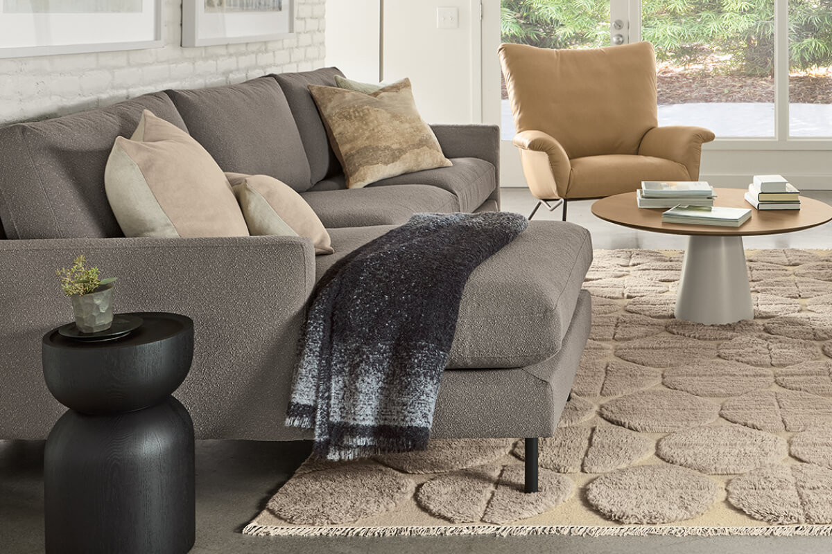 Grey Jasper sofa with chaise styled with pillows and throw beside a round pedestal coffee table and tan lounge chair.