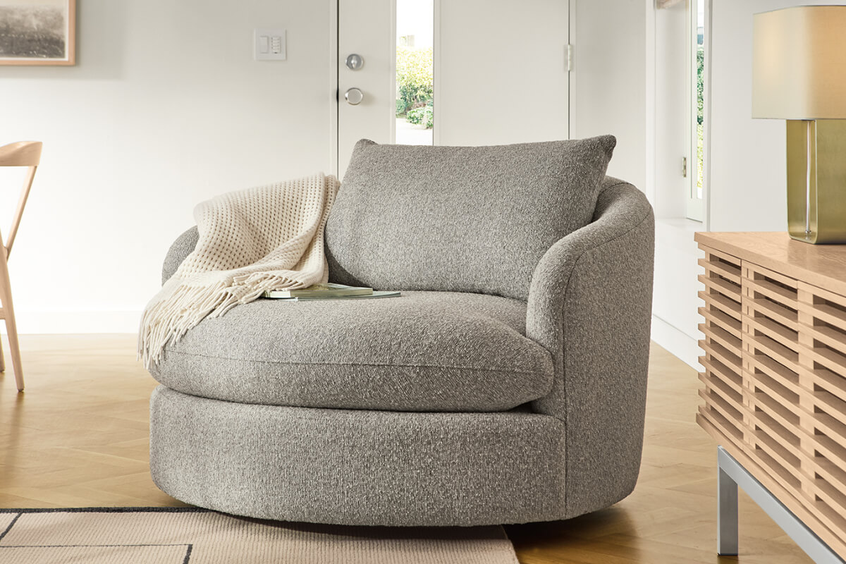 Grey Suri swivel chair with throw blanket beside a wood slat cabinet and lamp in a sunlit living room.