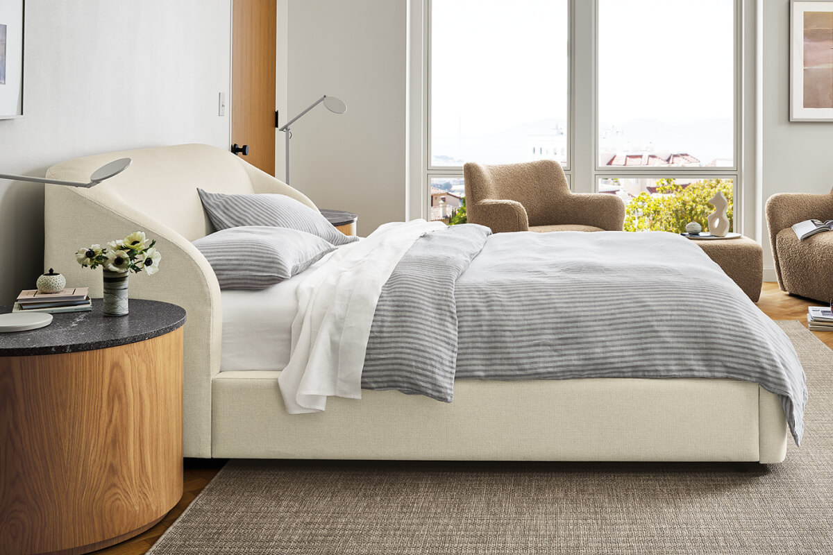Loxley upholstered bed in light fabric styled with grey striped bedding, pillows and a wood nightstand.