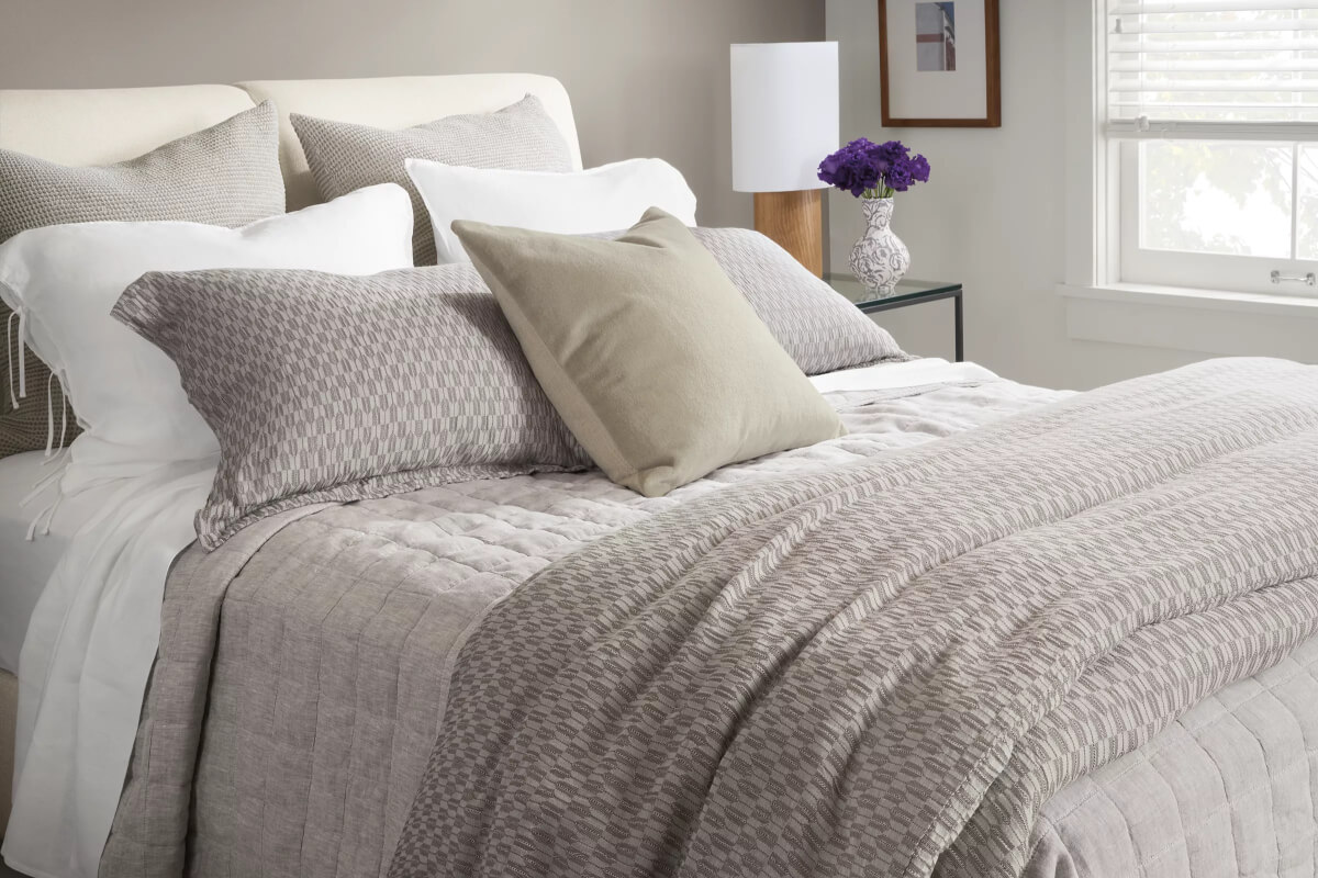 Grey Crofton bedding with neutral pillows and textured coverlet layered on a bed beside a nightstand and lamp.