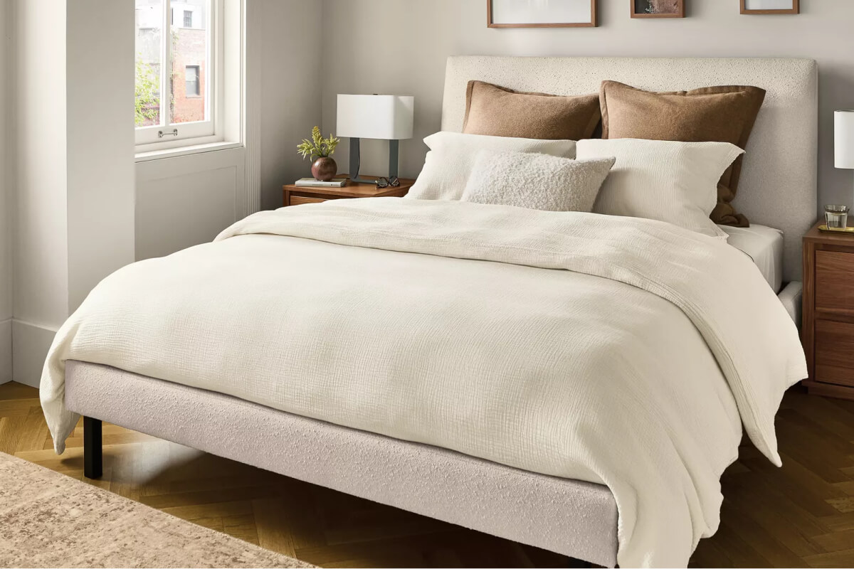 Ella upholstered bed in light fabric styled with white bedding and layered pillows beside a wood nightstand.