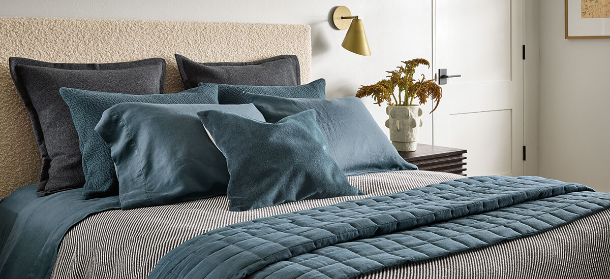 Quilted blue blanket and mixed-fabric pillows on bed with textured headboard and striped bedding.