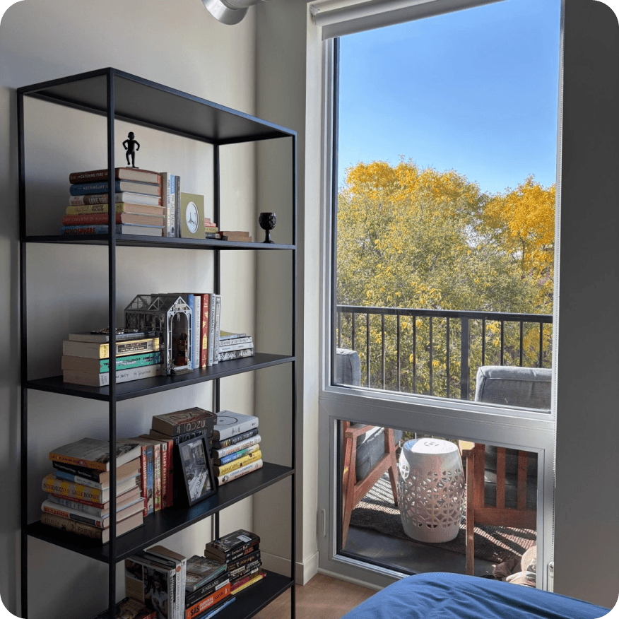 Slim bookcase with black metal frame styled with books and decor next to a window overlooking a balcony.