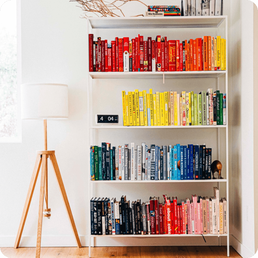 Slim white bookcase styled with rainbow-arranged books and decor next to a wood floor lamp.