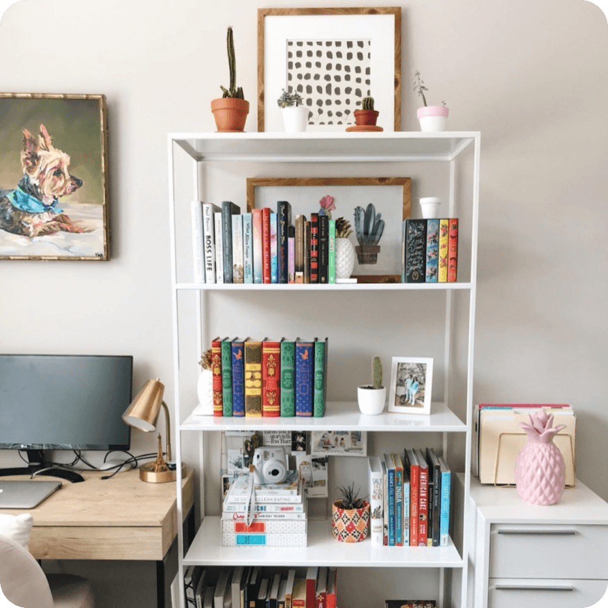 Slim white bookcase styled with books, framed art and small plants next to a desk setup.