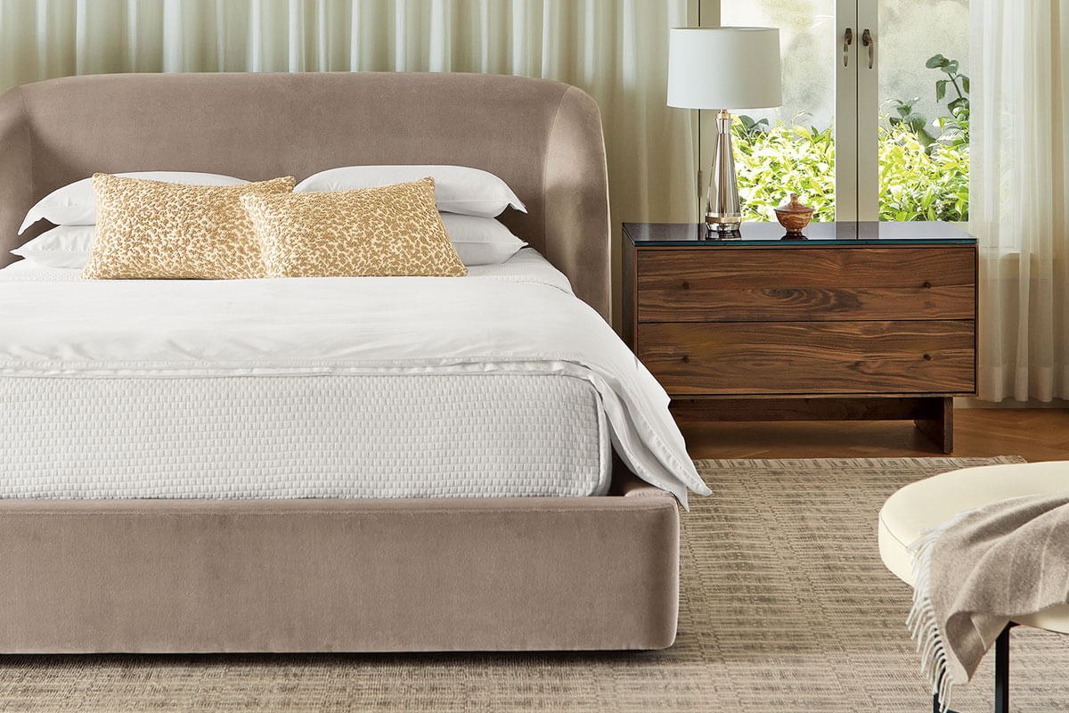 Loxley bed upholstered in beige with rounded headboard, paired with wood nightstand and table lamp.