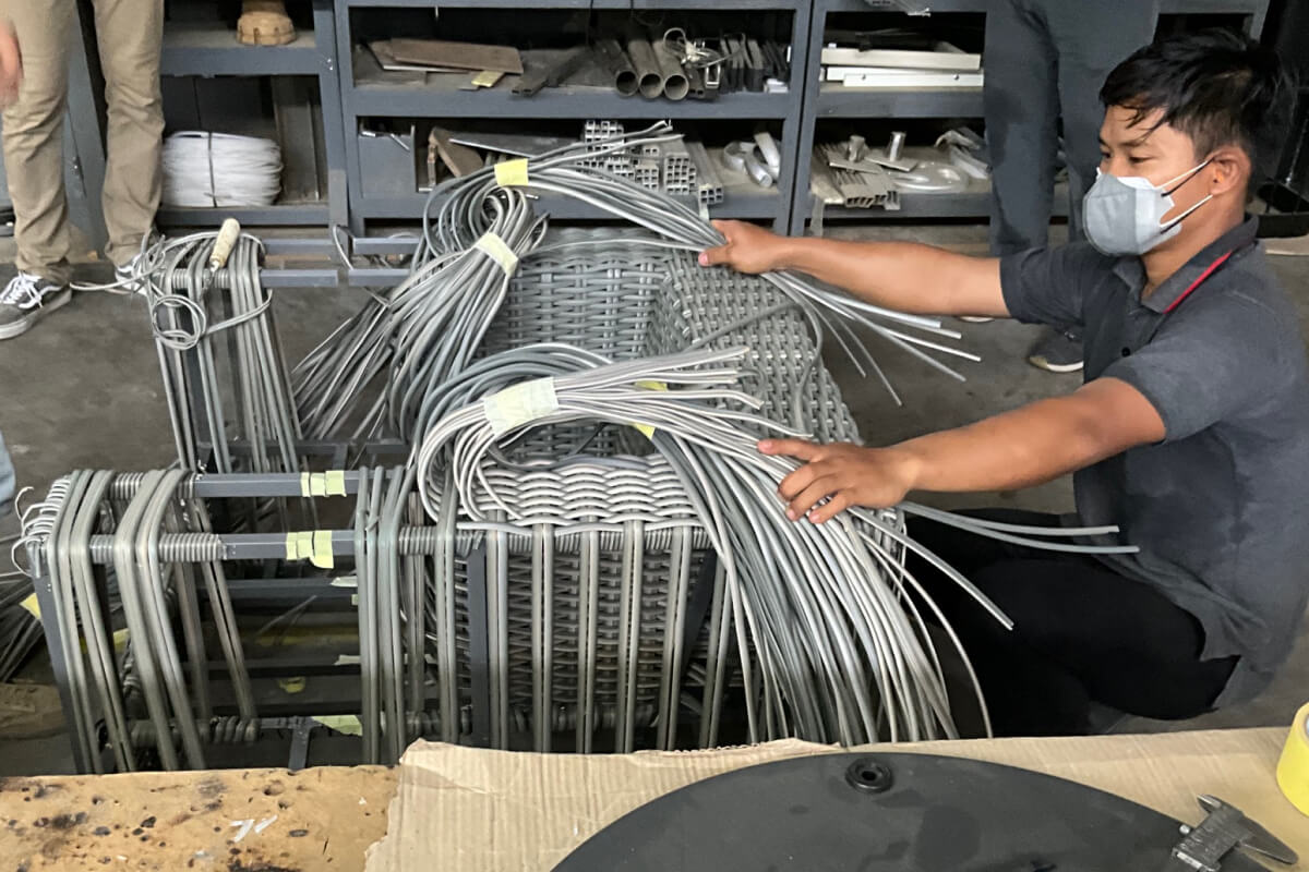 Worker assembling Madura seating with grey wicker strands over a frame, surrounded by stacked materials and tools.