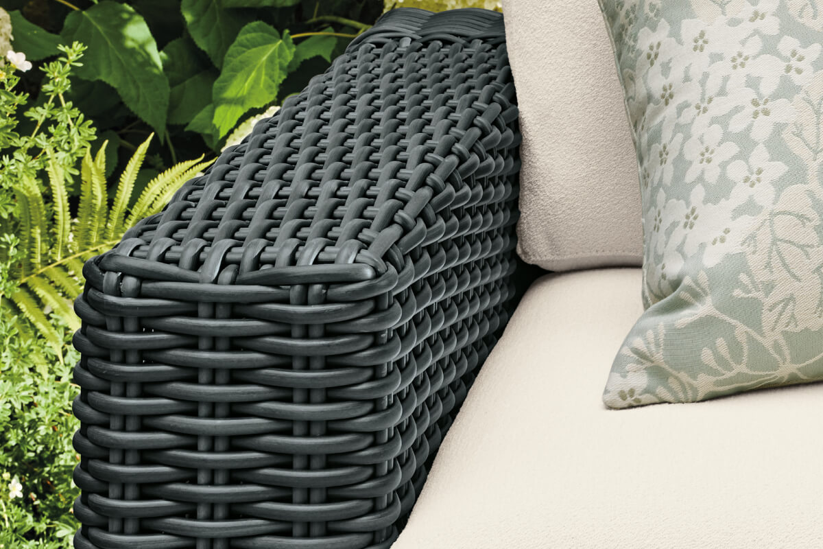 Madura seating detail showing dark grey woven wicker arm with a cream cushion and patterned pillow beside foliage.