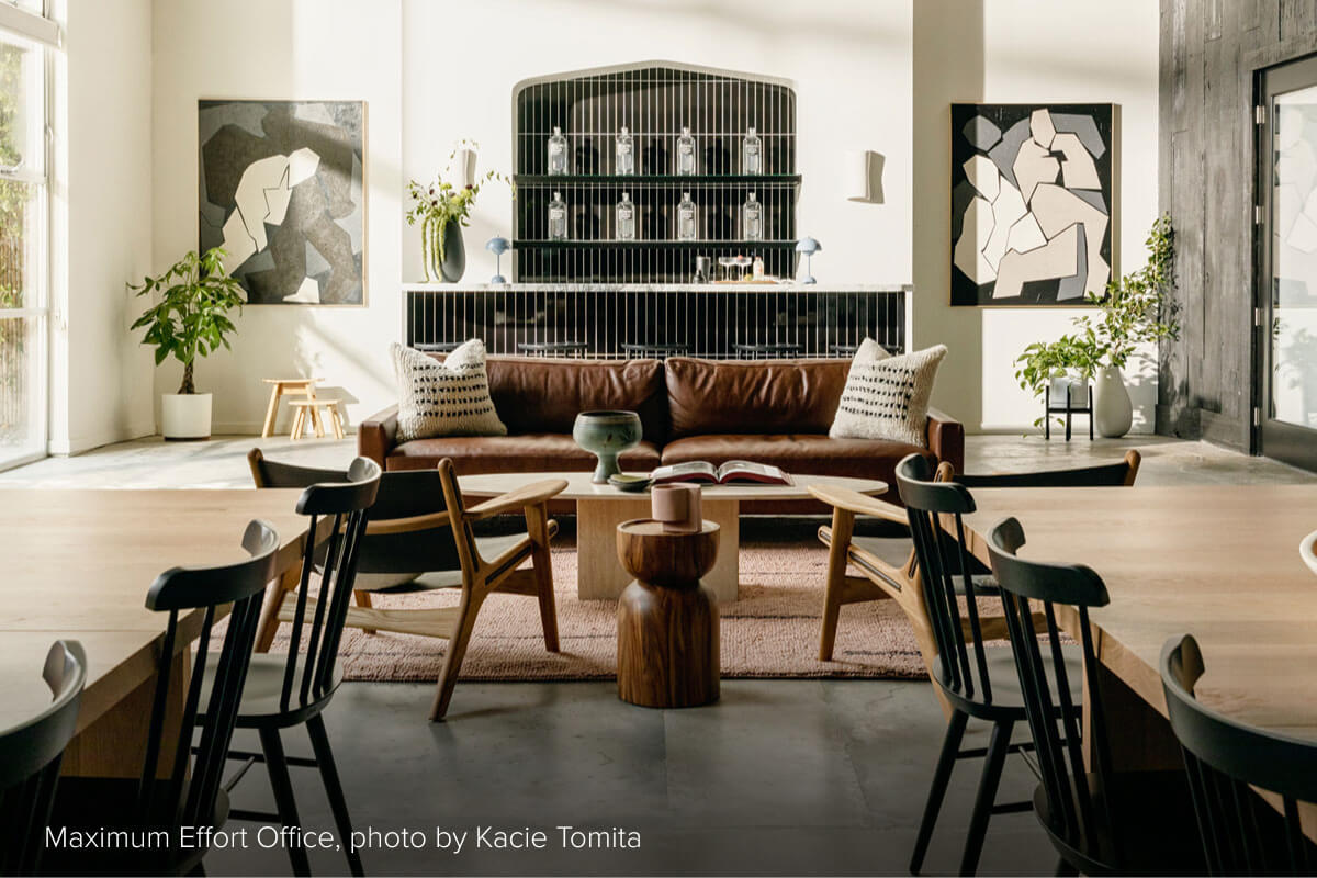 Brown leather sofa with two pillows behind a wood coffee table and stools, with dining tables in the foreground and a bar behind.
