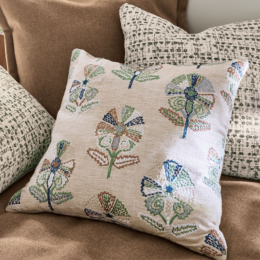 Wister pillow in cream fabric with embroidered floral pattern, layered with textured pillows on a brown sofa.