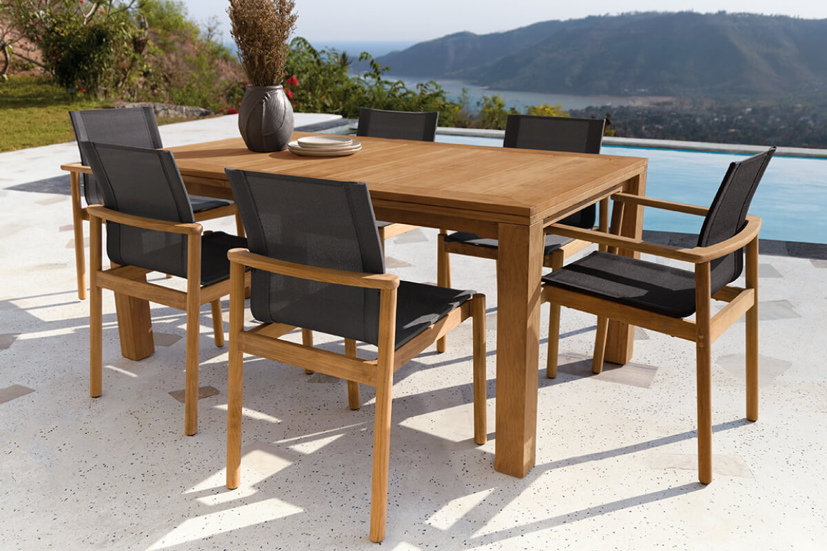 Capri table in teak with six chairs on a patio overlooking a pool and hillside landscape.