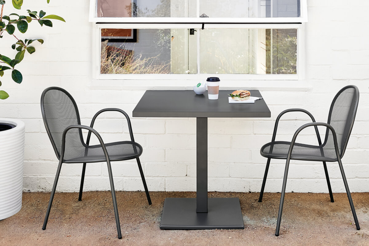 Matte black Maris table with Rio chairs on a patio against a white wall with window above.