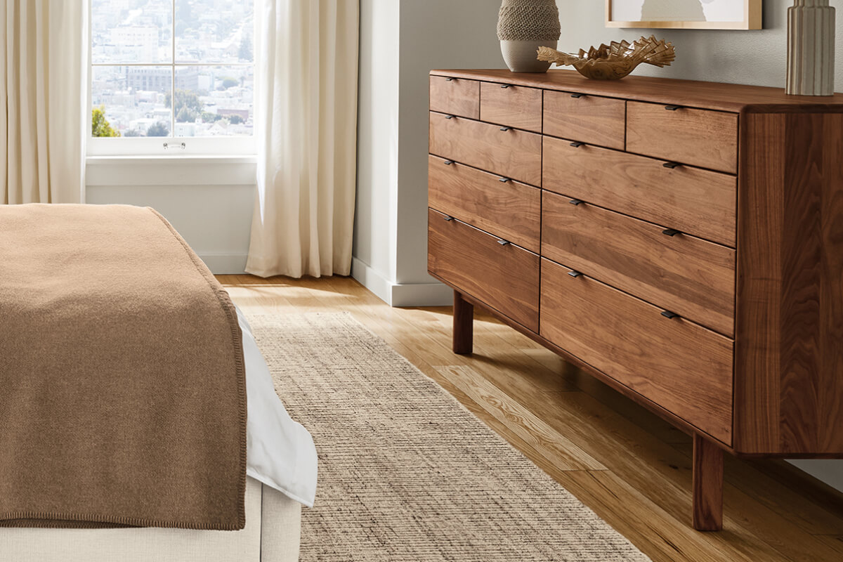 Hensley dresser in walnut with black pulls beside a bed, styled with decor near a window.