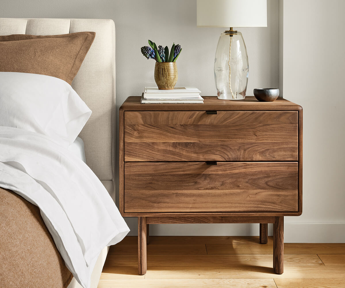 Hensley nightstand in walnut with two drawers beside a bed, styled with lamp and books.
